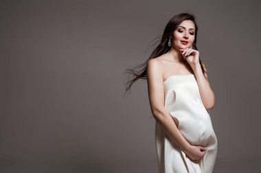 Mutlu Hamile Kadın. Hamile göbeği. Bebek bekleyen güzel hamile bir kadın. Annelik konsepti. Bebek partisi. Beyaz tekstil ürünü olan esmer kadın gride izole edilmiş. Saç üfleme. 30 haftalık hamilelik