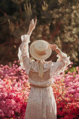 Hasır şapkalı kadın klasik filtre arka planlı çiçek açan çalılara bakıyor. Romantik kavram. Elbise giyen güzel bir kadın. Gün batımında doğada dinlenmek. Moda ve tarz. Yaz seyahati