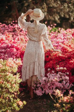 Hasır şapkalı kadın klasik filtre arka planlı çiçek açan çalılara bakıyor. Romantik kavram. Elbise giyen güzel bir kadın. Gün batımında doğada dinlenmek. Moda ve tarz. Yaz seyahati