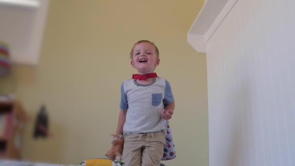 Niño pequeño con una capa salta sobre su cama — Vídeo de stock