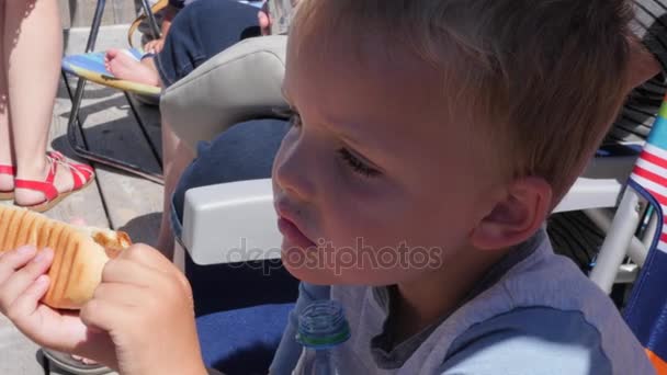 Хлопчик їсть хот-дог на прогулянці океаном — стокове відео