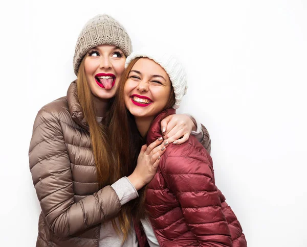 Närbild av mode porträtt av två glada vackra flickor vänner poserar för kameran inuti. Ljusa smink, hattar och vintern casual stil. Vit bakgrund, inte isolerade. Att ha roligt tillsammans. — Stockfoto