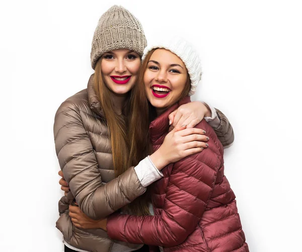 Módní portrét dvou veselých krásné dívky přátel pózuje pro kamery uvnitř zblízka. Světlý makeup, klobouky a zimní ležérní styl. Bílé pozadí, neizolované. — Stock fotografie