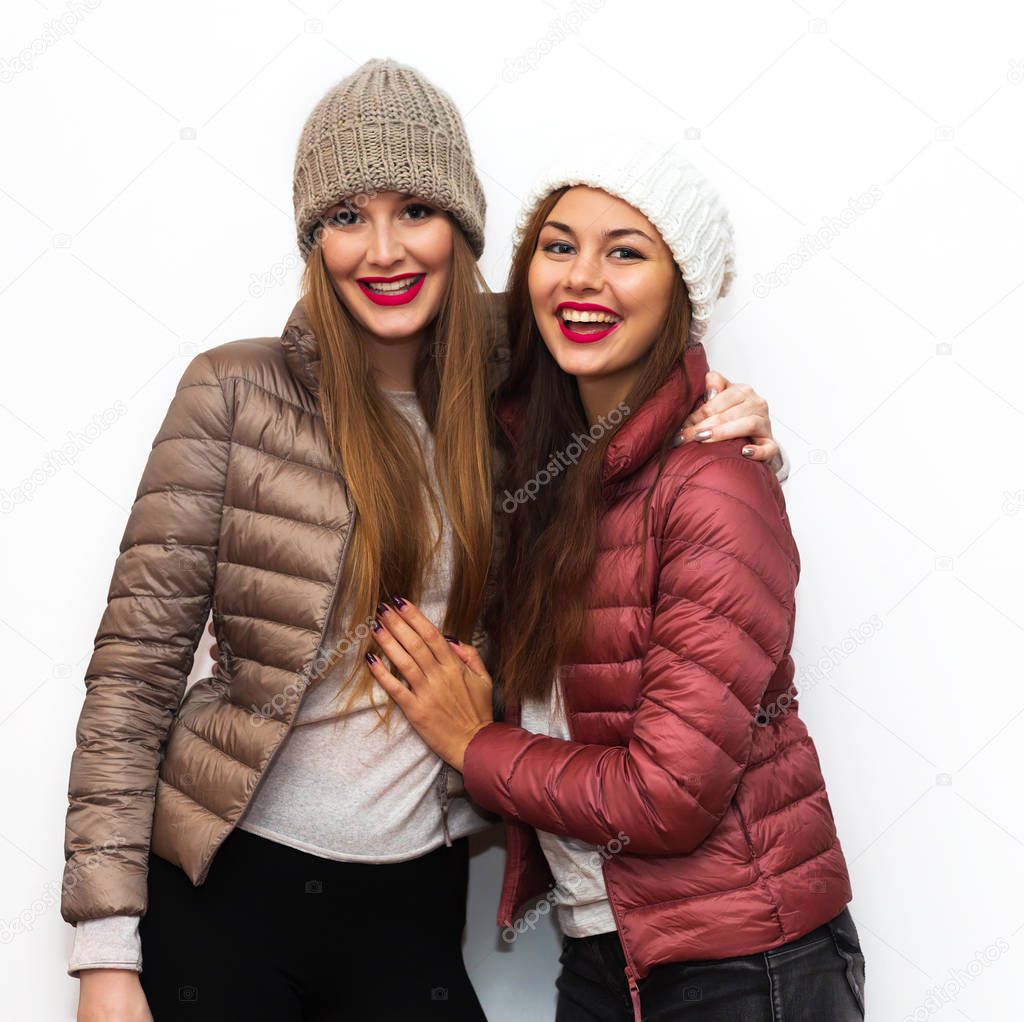 Close up fashion portrait of Two cheerful beautiful girls friends posing for camera inside. Bright makeup, hats and winter casual style. White background, not isolated.Having fun together.