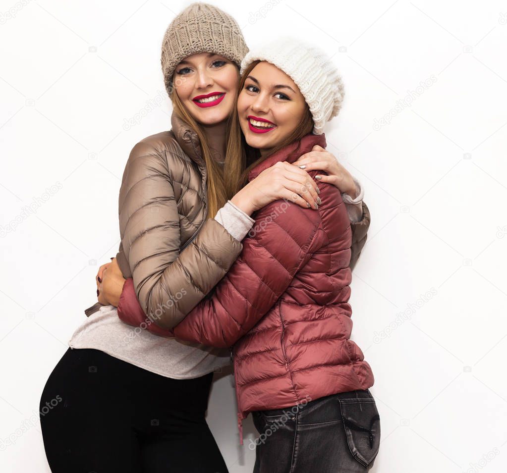 Close up fashion portrait of Two cheerful beautiful girls friends posing for camera inside. Bright makeup, hats and winter casual style. White background, not isolated.