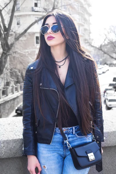 Mode portret van sensuele vrij stijlvol brunette meisje, trendy zwarte leren jas, vintage zonnebril, tas en blauwe jeanst dragen. Streetstyle. — Stockfoto