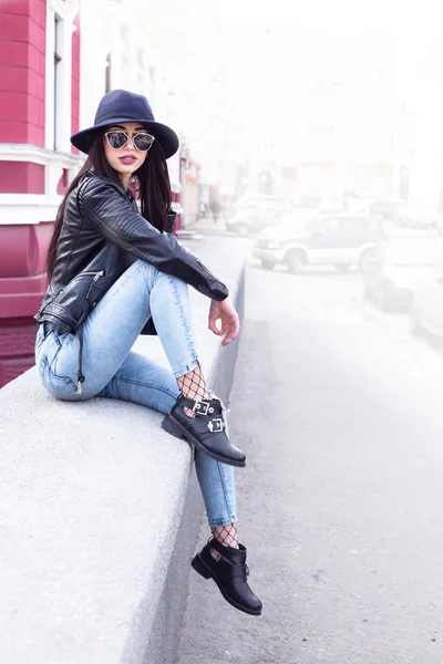 Concepto de moda callejera: retrato de una chica bonita, sentada en la calle afuera, con sombrero y chaqueta de cuero vintage, vaqueros azules, medias de rejilla, sombrero ancho, gafas de sol. . — Foto de Stock