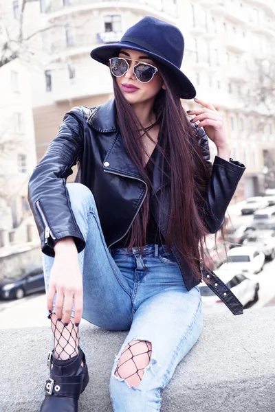 Concepto de moda callejera: retrato de una chica bonita, sentada en la calle afuera, con sombrero y chaqueta de cuero vintage, vaqueros azules, medias de rejilla, sombrero ancho, gafas de sol. . — Foto de Stock