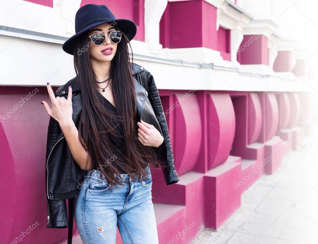 Closeup portrait of young beautiful fashionable woman with sunglasses. Lady posing on pink background. Model wearing stylish wide-brimmed hat and leather jacket. Girl looking at camera.Toned