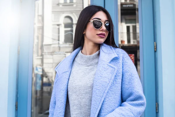 Concetto ultra fashion. giovane donna di moda indossa cappotto blu e posa su sfondo urbano di vetro blu. Stile street. donna d'affari. all'aperto. accessori alla moda . — Foto Stock