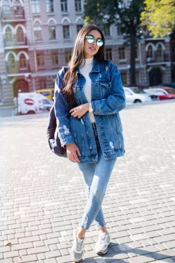 Hippi kız boş gri t-shirt, kot pantolon ve kaba sokak duvara, minimalist kentsel giyim tarzı poz sırt çantası giyiyor. Erkek kot pantolon ve uzun Kot ceket giymiş şık mutlu genç kadın