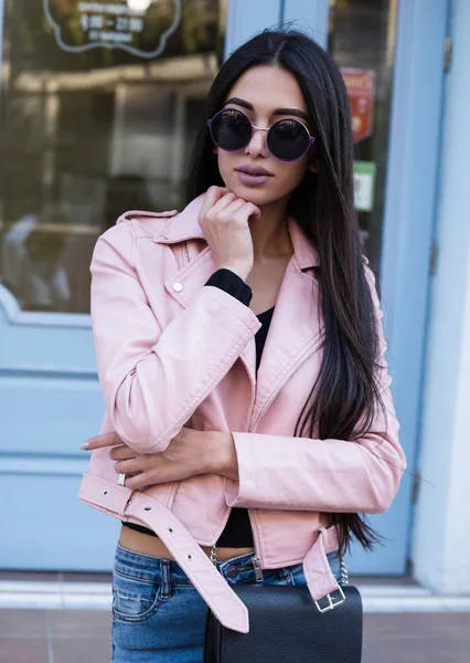 Street fashion concept - portret van een mooi meisje, wandelen in de straat buiten, het dragen van roze lederen jas, grijze trui, grote roze zak, zonnebril. Zachte warme vintage kleurtoon. Zonnig weer. — Stockfoto