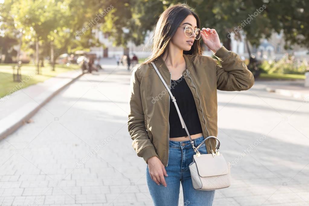 Fashion close up portrait stylish woman in fall spring casual outfit walking in city. Street style.young beautiful woman in street background in khaki bomber coat,jeans and bag,autumn fashion trend.