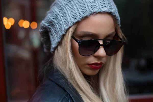 Primer plano retrato femenino de modelo de moda con el pelo largo rubio rizado con gafas de sol sentado y posando al aire libre. Jeans, gafas de sol, sombrero, chaqueta. Joven mujer con estilo — Foto de Stock