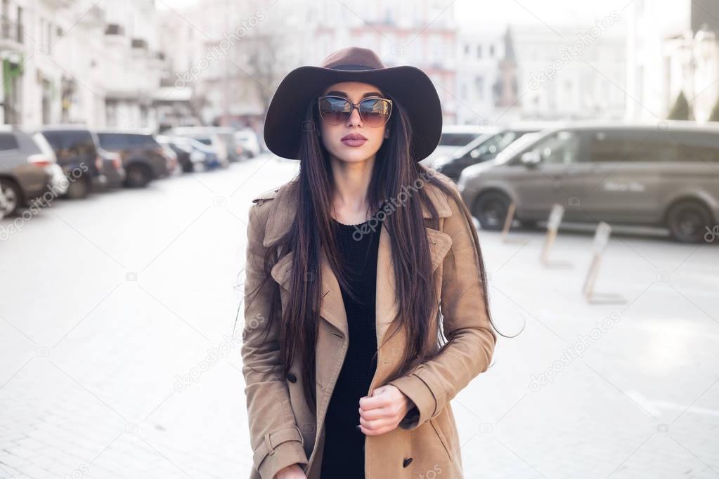 Outdoor portrait of young beautiful fashionable woman with long hair walking in the street. Model looking aside, wearing stylish classic beige coat, hat, accessories. Female fashion, beauty concept