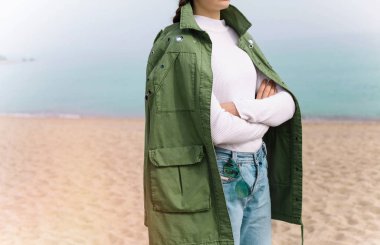 Tanınmayan modeli rahat kıyafet uzunluğundaki haki ceket, kot pantolon, beyaz kazak giyiyor. giyim moda minimalist tarzı. Bahar ve sonbaharda sezon okyanus arka plan, avuç içi ile plaj, sokak moda.