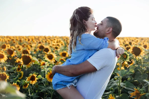 Shot of young woman being carried by her boyfriend in sunflower field. Couple having fun on their summer holiday.handsome guy with a beard in a blue denim shirt gentle hugs, hand holding and kissing