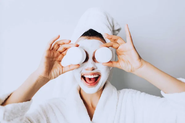 woman has fun with a facial mask.Young girl with facial mask, holding bottles of cream  looking at camera over white background. Cosmetic procedure. Beauty spa and cosmetology.Spa Facial Mask. Spa day