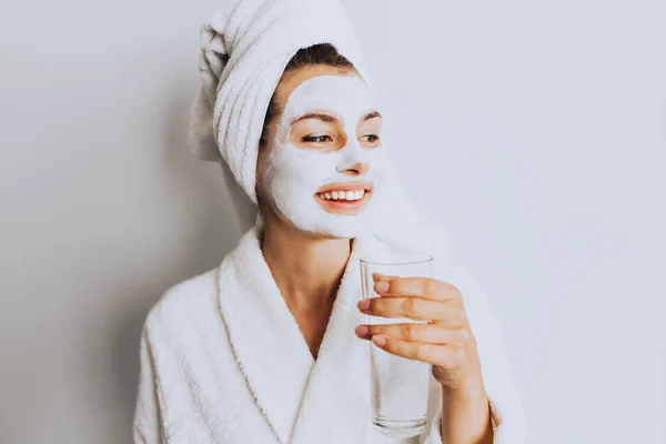 Face and body skin care, face treatment at home concept, wearing bathrobe and towel. Beautiful young woman drinking a fresh glass of water at home