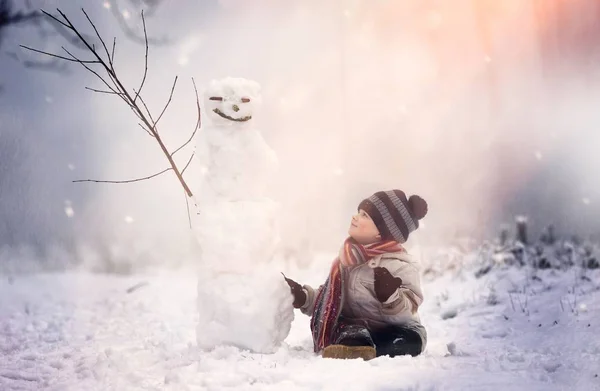 Niño sentado cerca de muñeco de nieve en el paisaje de invierno . — Foto de Stock