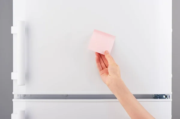 Mano femminile che tiene nota di carta rosa sul frigorifero bianco — Foto Stock