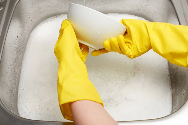 Hands in gloves washing white bowl under running tap water