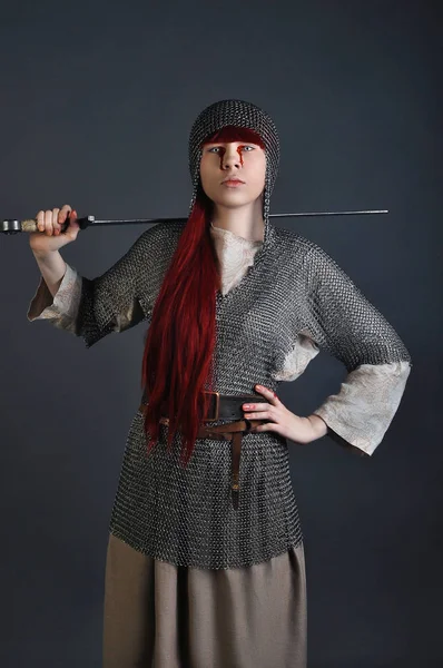 Medieval girl in chain mail with a sword with red hair stands on a gray background with blood on her hands