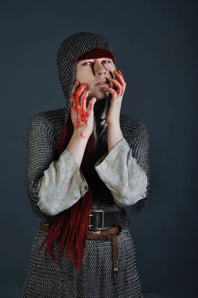 Medieval girl in chain mail with red hair stands on a gray background with blood on her hands