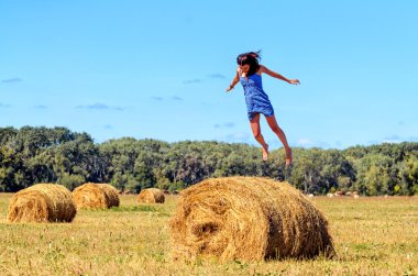 Genç kadın bir alanda haystack atlar. Elleri ayrı. Mavi gökyüzü ve arka plan, yeşil ağaçlar. Büyük demetleri saman sarı çim yalan. Fotoğraf seyahat. Kız yalnız