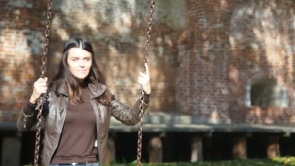 Beautiful woman is having fun swining swing underneath a tree in nature. Shadows of branches casts to an old red break wall on background. Girl wears blue jeans and brown jacket — Stock Video