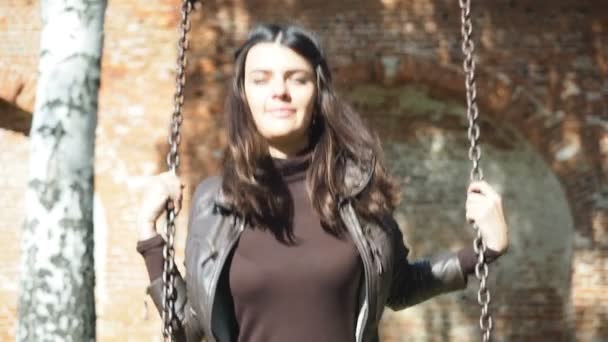 Bella donna si sta divertendo swining swing sotto un albero in natura. Le ombre di rami lanciano a una parete di pausa rossa vecchia su sfondo. Ragazza indossa jeans blu e giacca marrone — Video Stock