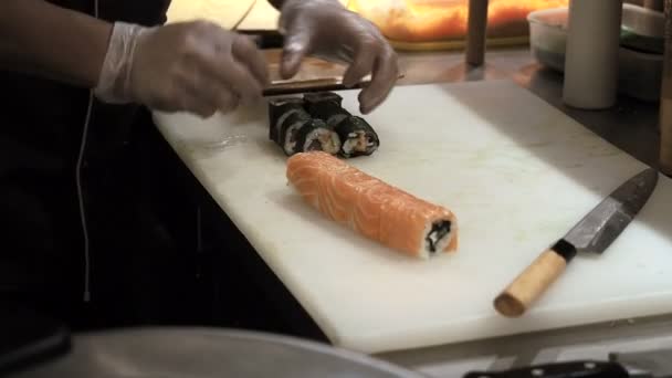 Process of cutting orange sushi rolls by knife. Man rolling up sushi set using bamboo mat. Prepared sushi rolls pass through on foreground — Αρχείο Βίντεο
