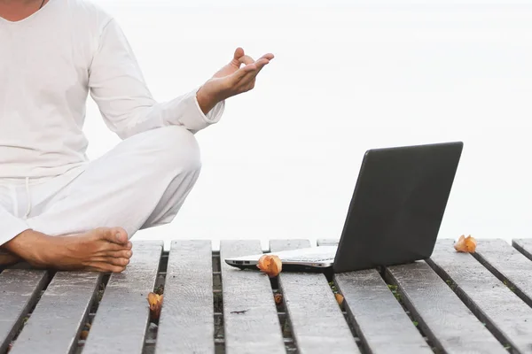 Hombre en ropa blanca meditando yoga con portátil en muelle de madera — Foto de Stock