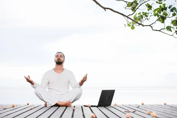 Homem de roupas brancas meditando ioga com laptop no cais de madeira — Fotografia de Stock