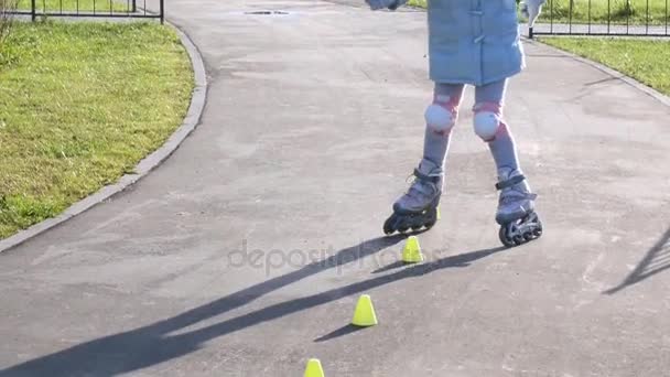 Маленькая девочка учится кататься на роликах между конусами в парке — стоковое видео