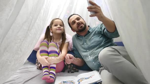 Отец с веселой маленькой дочкой играет со смартфоном и разговаривает по скайпу по мобильному — стоковое видео