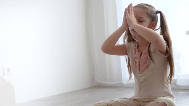 Niña linda sentada en la pose de loto de yoga y medita cerca de la ventana — Vídeos de Stock