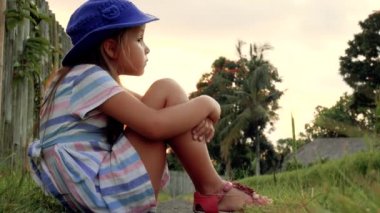 Çimenlerin üzerinde oturan ve yaz bahçede oynarken mutlu küçük kız. Açık havada. Günbatımı.