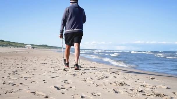 Stary człowiek używa kijów do skandynawskiego spaceru po plaży morskiej, słoneczny dzień — Wideo stockowe