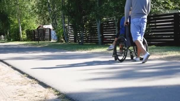 Kesenangan keluarga. Anak muda dengan ibu cacat atau f di kursi roda berjalan di taman — Stok Video