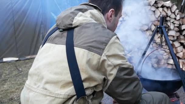 Dos hombres con ropa militar cocinando al aire libre en caldero de hierro fundido. Cocinar en un fuego. La comida en el caldero sobre el fuego en la naturaleza — Vídeo de stock