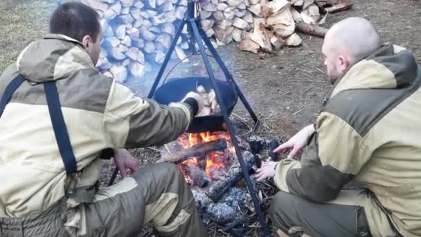 Dos hombres con ropa militar cocinando al aire libre en caldero de hierro fundido. Cocinar en un fuego. La comida en el caldero sobre el fuego en la naturaleza — Vídeos de Stock