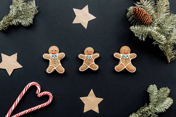 Three gigngerbread men on a black backdrop surrounded by papr stars, small fir tree branches, a cone and two candy canes