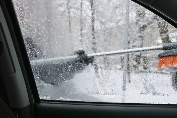 A man cleans a car from snow. Winter car care. Inside view of the car. Blurred background behind the glass.