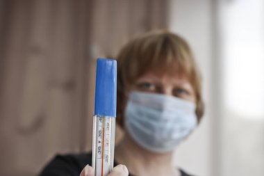 Ön plandaki eldeki vücut sıcaklığını ölçmek için tıbbi termometre, arka plan bulanık. Bir kadın elinde bir termometre tutuyor. Yüksek vücut sıcaklığı hastalığın işaretidir..