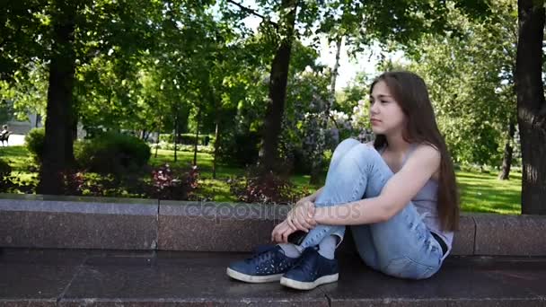 Aranyos tizenéves lány ellenőrzése telefon és a várakozó hívás ül a parkban. Videofelvétel Hd felvétel statikus kamera. — Stock videók