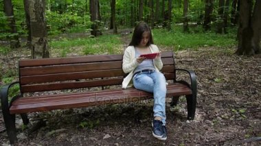 Güzel genç kız tablet bilgisayar ile parkta bankta oturur. Statik kamera film.