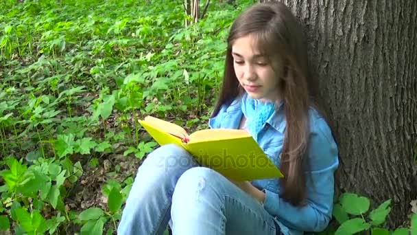 Portrait d'adolescente mignonne lisant le livre et tournant la page appuyé contre le tronc d'arbre dans la forêt au printemps, étudiant en plein air . — Video