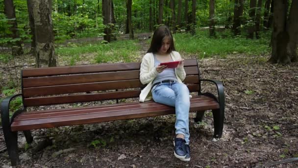 Güzel genç kız tablet bilgisayar ile parkta bankta oturur. Statik kamera film. — Stok video