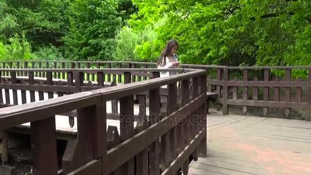 Teenager girl using mobile phone on the wooden bridge. Young teen texting message on smartphone walking. Motion camera, panorama with steadicam. — Stock Video